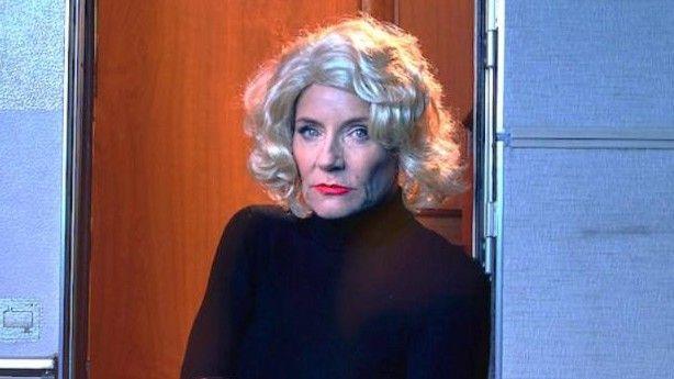 The actress Michelle Collins in a Marilyn Monroe styled blonde curly wig and black polo neck and leggings with white stilleto shoes sits on the steps of a rather grubby caravan. She looks very angry.
