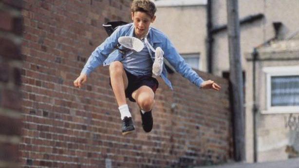 A scene from the film Billy Elliot shows him up in the air while he is mid jump on a street next to a brown brick wall. He is wearing black shorts and shoes, white socks and a blue shirt over a blue and white striped t-shirt. A pair of white trainers in hanging around his neck.