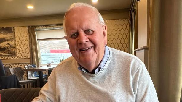 Photograph of Phil Woodford, who is smiling while sitting in a tearoom. He has grey hair and is wearing a pale grey jumper over a light blue shirt.