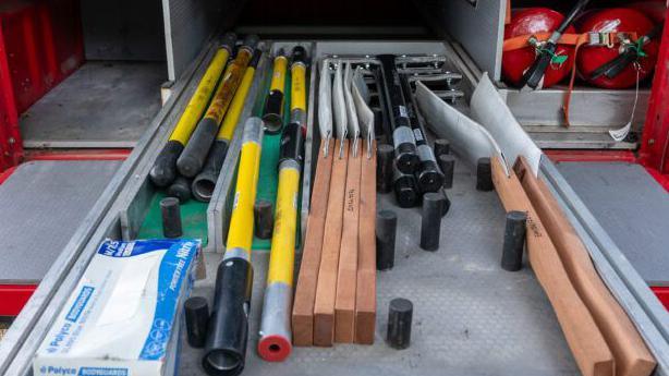 Different equipment, including items that look like long shovels, are in rows on a sliding compartment in the back of a vehicle. There are also two red fire hydrants on the right.