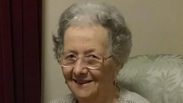 A woman smiling while looking straight at the camera. She is sitting in a cream armchair and is wearing a cream knitted jumper and glasses.