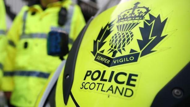 An image of 'POLICE SCOTLAND' lettering in bold, on a yellow police vehicle, with an officer dressed in yellow luminous uniform in the background.