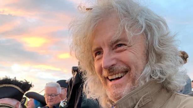 Paul Manning with beard and long hair smiling in front of crowd and sunrise.