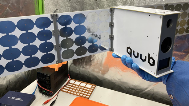 A small, white satellite with QUUB branding on is being held in a person's hand. They have blue gloves on. Other equipment is below.