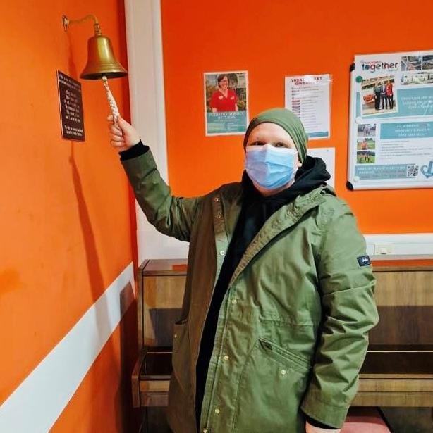 Christy Esslemont, woman wearing green jacket, and face mask and head scarf,  ringing the 'free from cancer' bell, in an orange room