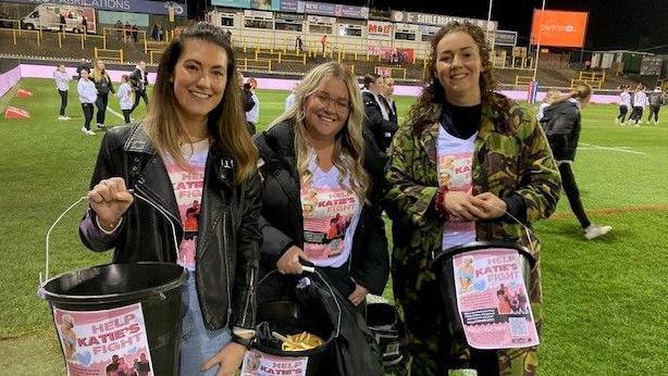 Three women hold buckets aloft, all of them bearing a pink-coloured poster headed 'Help Katie's Fight'. The women are all wearing t-shirts with the same slogan and details on. They are stood on a floodlit sports pitch while children walk in the background.