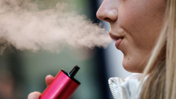 A close up shows the bottom of a woman's face - she is blowing out vapour and holding a pink/red vape in her hand