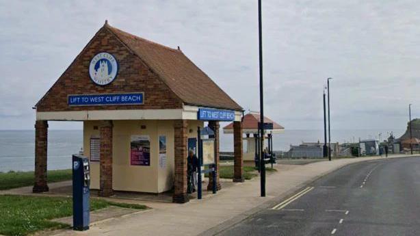 An open sided building stands next to a parking meter. There are two blue signs saying 'Lift to West Cliff beach', with 'West Cliff Whitby' above one of them, around a silhouette of a building. There are also posters on the inner yellow walls. It is next to a road.