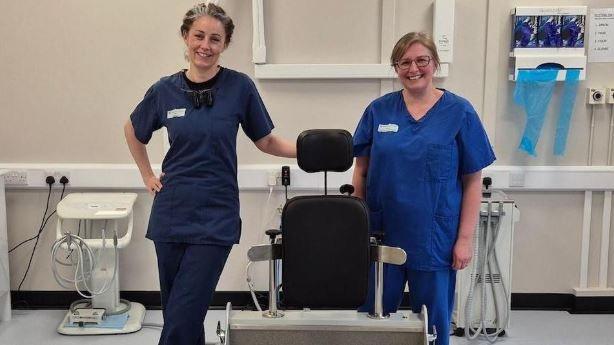 Dentists Beverley McClung, who wears a navy uniform, and Patricia Newson, who wears a lighter blue uniform, stand next to the diaco ramp, which holds a wheelchair and has the top part of a dentist chair attached.