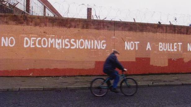 A wall painted orange with barbed wire on top of it and white writing in capitals across is saying 'NO DECOMMISSIONING NOT A BULLET'. There is a man cycling past on a blue bike in dark clothes and a hat.