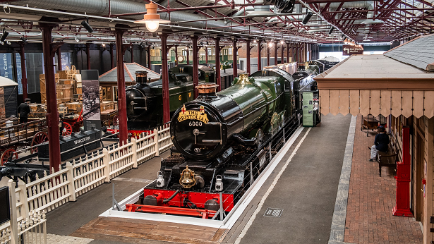 Green and black steam trains in a museum. There stationary at platforms with people sitting at the side.