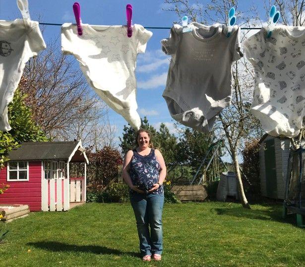 Jacqui, who is heavily pregnant, is wearing blue jeans and a blue vest top standing in a garden with a washing line in the foreground. The washing line has baby clothes pegged to it.