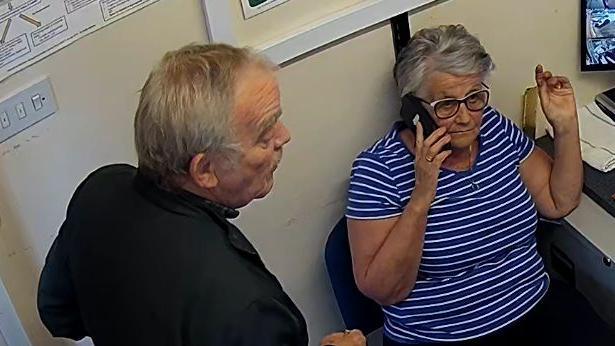 A CCTV image of a man and a woman in an office room. The man is wearing a black shirt and is stood facing the woman, who is wearing a blue shirt with stripes as she holds a mobile phone up to her ear.