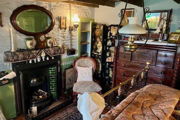 The image shows a room inside the house. There are various period pieces, including a mirror in an oval frame, a chair, wooden chest of drawers and a fireplace.