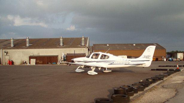 Turweston Aerodrome showing single-storey hanger building to the left with four tall metal chimneys and two large doors wide enough for planes. There is a further brown-coloured building adjacent. In front of this is a white light plane parked on the tarmacadam.