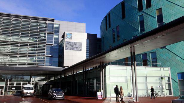 The Royal Victoria Infirmary in Newcastle. It is a tall grey building, with a circular green building next to it. There is an ambulance and a car parked outside. A few people are standing near the entrance. 