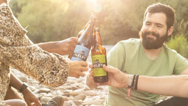 Four people sit on a pebble beach by a river, clinking their bottles of Ilkley Brewery beer