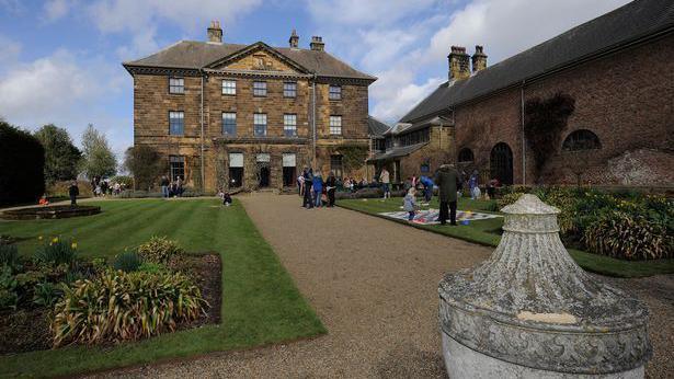 A view of Ormesby Hall from a gravelled footpath leading up to the stone building. A stone urn stands in the foreground and families can be seen on the grass leading up to the hall.