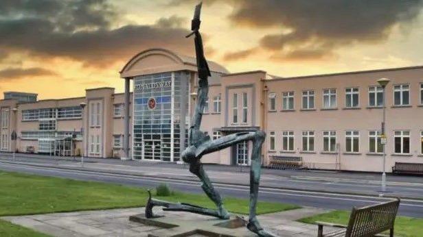 The Isle of Man Airport building at dusk. It is a long cream-coloured building with a large glass front that has Ronaldsway written above it and a road running along side it. There is a sculpture of the Three Legs of Man on the grass in the fore.
