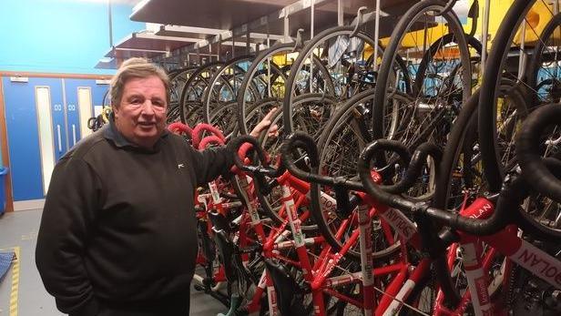 Alan Davis who has brown hair and is wearing a black jumper, standing next to a rack of red, white and black racing bikes