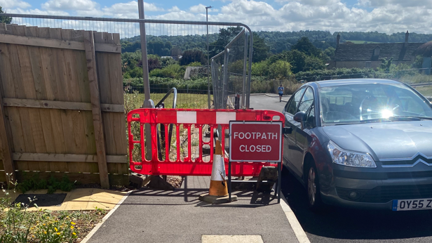 A red fence with an orange cone and a sign reading "footpath closed" sits in front of a stretch of pavement.