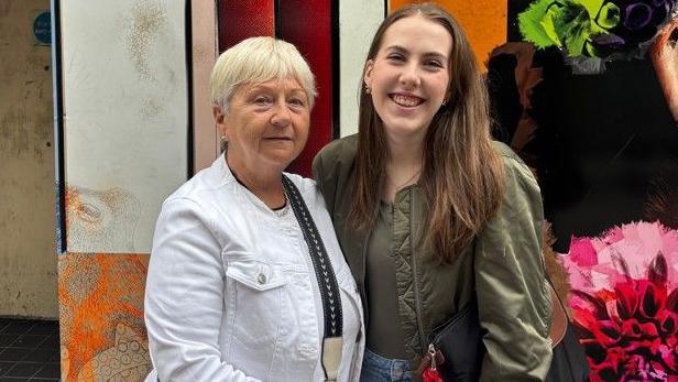 On the left is Agnes, wearing a white jacket, black jeans with a black and cream cross body bag. She has short blonde hair. Beside her on the right is her granddaughter Abbie, wearing blue jeans, a khaki bomber jacket with a black shoulder bag.