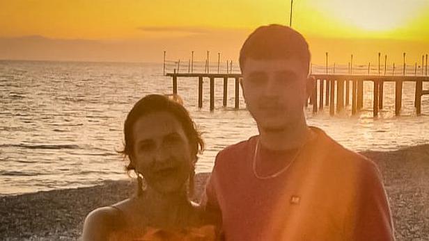 Rachel and her son stand arm in arm on the beach wit the sun setting in the background behind a pier. 