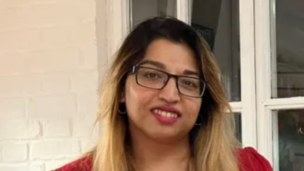A close-up image of Lowestoft Mayor Nasima Begum. She is wearing glasses and a red t-shirt and is standing in front of a window. She is smiling into the camera.