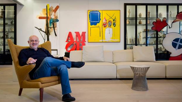 Ahmad sits on an armchair with his legs crossed in his penthouse in Beirut. He is wearing jeans and a dark shirt. Behind him is a wall of art and a cabinet with objects in. There is a cream sofa against the wall.