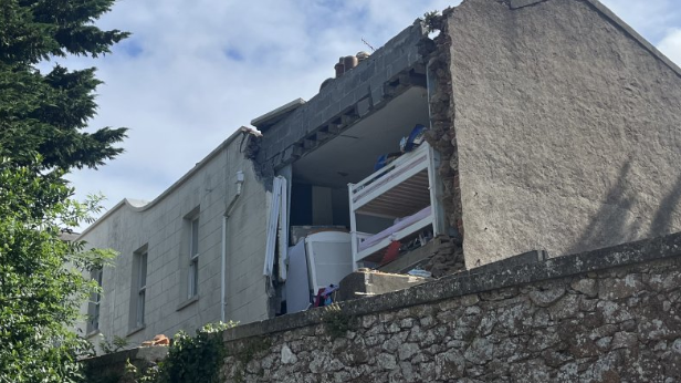 The damaged building with a wall missing and furniture including a bunk bed visible