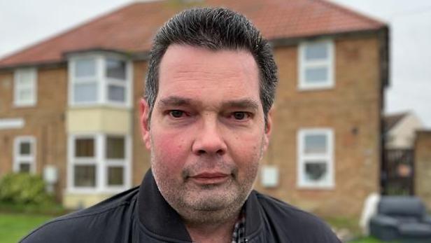 Phillip Shaw smiles at the camera. He has dark, short hair. He is wearing a black coat with a checked shirt underneath. He is standing in front of a house, which is out of focus.