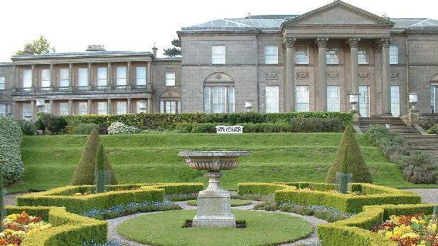 Plants and trees in a formally designed garden in front of the neo-classical facade and pillars of Tatton Hall mansion.