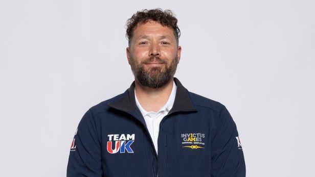 Daniel Bennet is wearing a navy blue Invictus Games Team UK zip jacket. He has his arms down by his side and is smiling at the camera. 