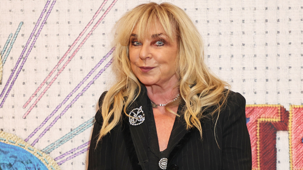 Helen is stood in front of a colourful backdrop. She has long blonde hair and is wearing a black suit jacket and smiling at the camera.