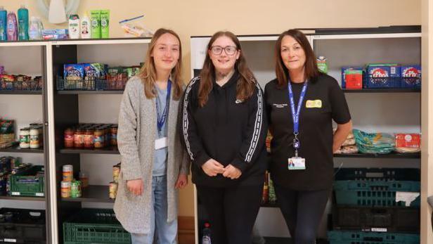 Rachel Fairless is standing in the middle of the pantry with two other women. Behind them are shelves filled with pantry food and tins.