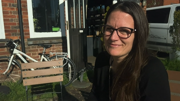 A woman with straight brown hair and black glasses sits outside a brick building