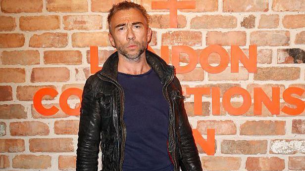 James Holder is standing in front of a brick wall with orange lettering on it. He is wearing a black leather jacket and a navy t-shirt.
