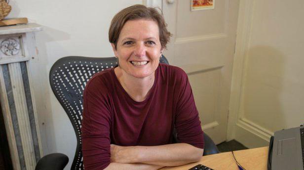 Helen Bedser from Julian House sits smiling at an office desk. She is wearing a dark red long-sleeved top and is sitting on a modern-style office chair