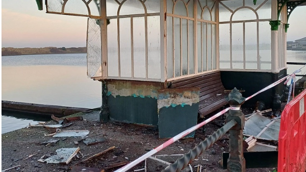 A Victorian style shelter next to a body of water which has been partially smashed, debris including pieces of wood and glass is scattered on the ground.