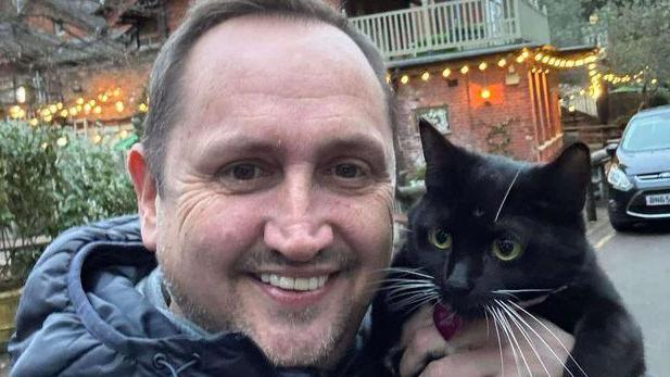 A man smiling and looking at the camera while holding a black cat with green eyes.