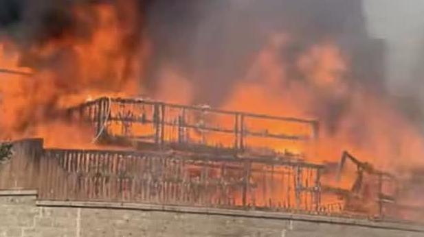 A burning fence and the frame of a house is all that is visible in this picture from below a wall of orange flames. Black smoke is coming up from a bungalow above a grey brick wall.
