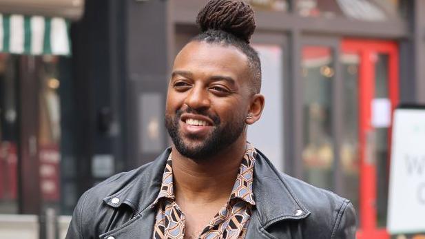 Oritse Williams, with his black hair in a top knot, is wearing a black, orange and blue patterned shirt and a black leather jacket