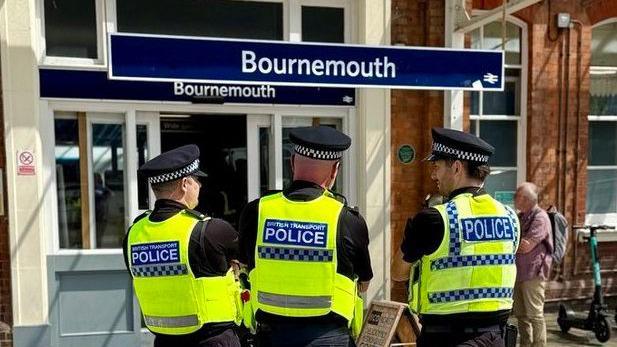 Three male police officers stand with their backs to the camera looking towards Bournemouth train station. There is a large blue sign for Bournemouth in front of them, marking the station entrance. All three officers have their arms crossed. They are wearing black short sleeved t-shirts, with black peaked caps which have black and white check around them and high-vis yellow vests. The vests have a blue rectangle on the back and white writing which says: "British Transport Police".