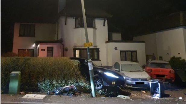 A white house in front of which there are three cars, two of them, which are white and red, are parked in a driveway and a third black car seen behind a hedge next to a wonky lamp-post. The black car has its lights on and there is a knocked over bin and other rubbish nearby.