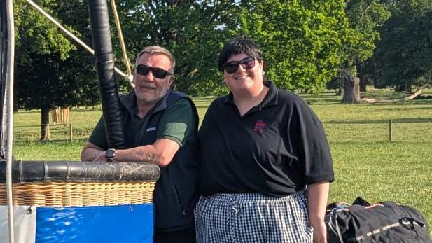 Lorna Hankey wearing a black polo top and checked trousers is standing next to a balloonist who is wearing a dark green polo top and blue gilet. The pair of them are standing next to a balloon basket.