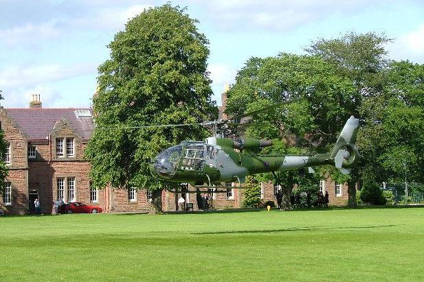 A camouflaged helicopter hovers above a large grassy lawn. Behind it are large trees and a red-brick building.