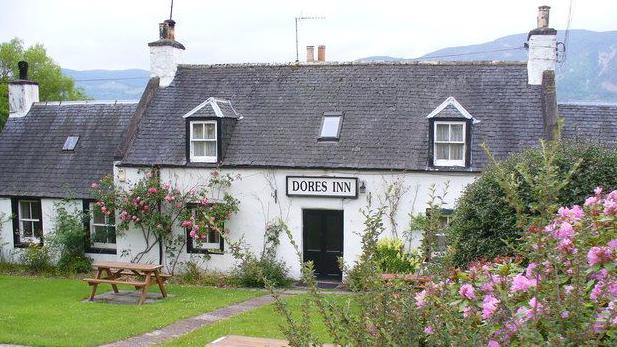 The white-walled two-storey building has a garden in front of it with shrubs with pink flowers.