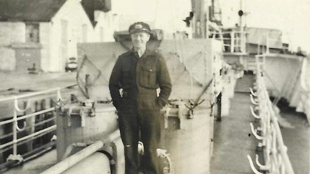 Old black and white photograph of a sailor. He is wearing a dark and unadorned uniform and a cap with a badge on the front. He is standing, hands in pocket, on the deck of a ship.