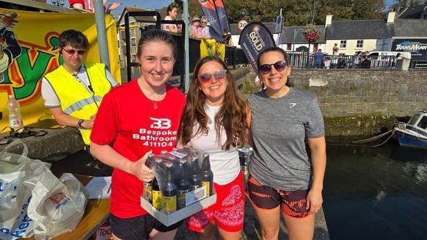 Three women in brightly coloured shorts and t-shirts smile at the camera. Abigail Quinn, the winner, stands in the centre. One woman holds a case of beer in glass bottles. They stand by Castletown Harbour in the sun.