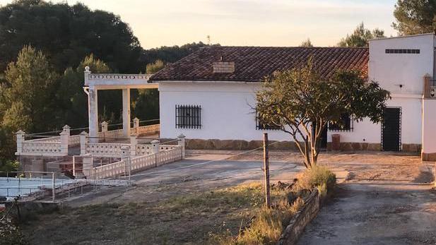 A Spanish country house. It is detached and painted white with barred windows and a tiled roof. There is a terrace to the left with decorative balustrades and open land and a tree to the right.